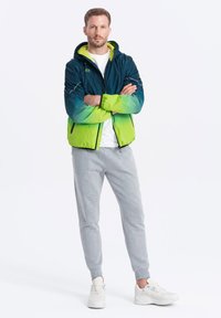 Herrarnas huppjacka i en blå till grön gradient, med dragkedjefickor och elastiska manschetter, i kombination med gråa sweatshirtbyxor och vita sneakers.