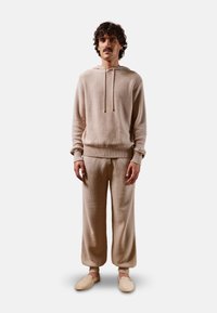 Hoodie en tricot beige avec un motif texturé, capuche à cordon, et poignets côtelés, associé à un pantalon ample assorti et des chaussures à enfiler.