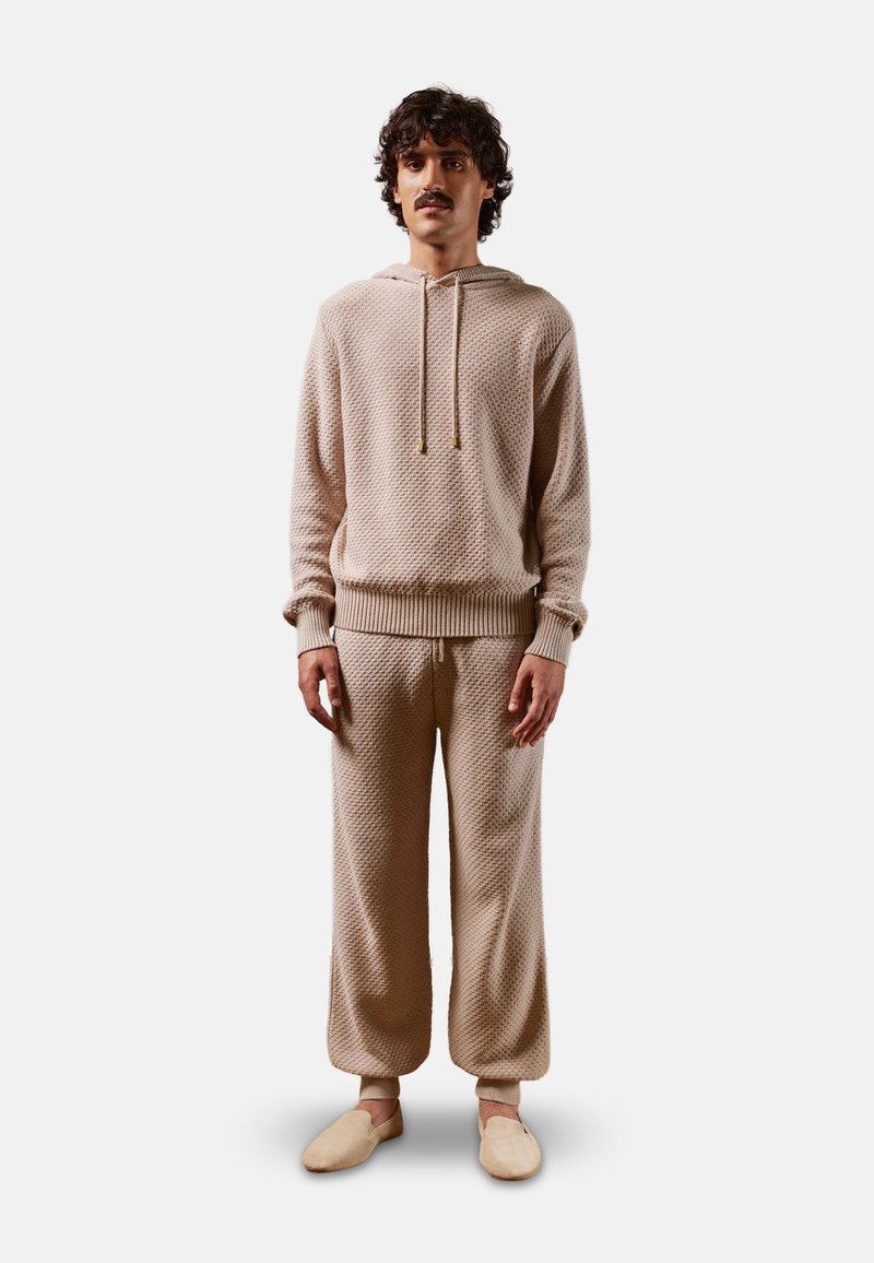 Hoodie en tricot beige avec un motif texturé, capuche à cordon, et poignets côtelés, associé à un pantalon ample assorti et des chaussures à enfiler.