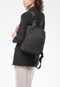 Zaino nero con tasca frontale in pelle texturizzata, hardware dorato, doppi manici superiori e tracolla regolabile. Corpo in tessuto liscio.