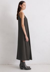 Dunkelgrünes ärmelloses Kleid aus glattem Stoff, mit einer lockeren Passform, die bis zu den Knöcheln reicht, kombiniert mit schwarzen Stiefeletten.