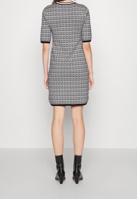 Femme portant une robe courte à manches courtes, longueur genou, avec un imprimé répétitif "DKNY" noir et blanc sur l'ensemble, et des bottines noires.