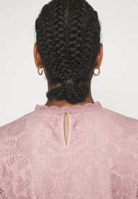 Blusa in pizzo rosa chiaro con intricate fantasie floreali, colletto scallopato e dettaglio a goccia sul retro. Presenta un tessuto testurizzato.
