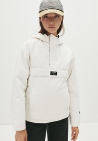 Hvid isoleret jakke med høj krave, frontlomme og justerbar hætte. Parret med en grå og beige kasket. Blød tekstur, moderne design.