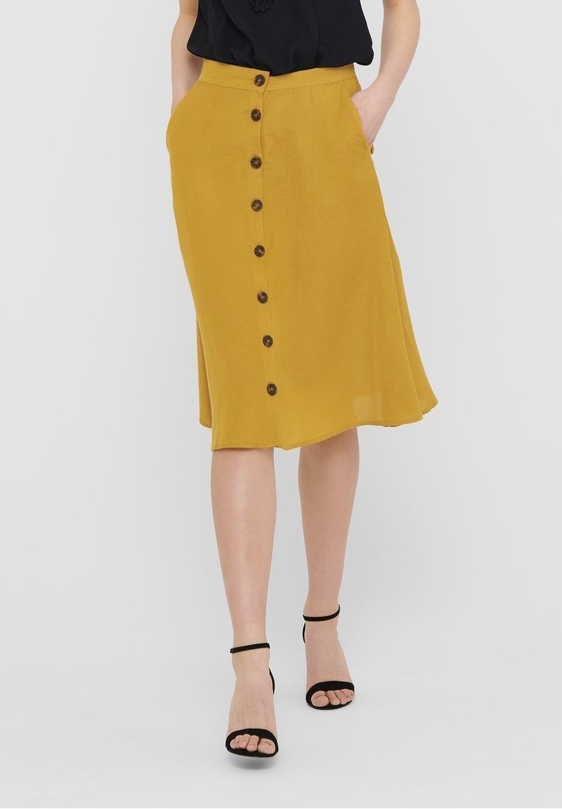 Femme portant une jupe midi moutarde boutonnée à l'avant avec des poches, associée à un haut noir sans manches et des talons noirs à bride cheville.