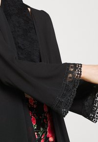 Svart kimono-liknande jacka med spetstrim på ärmarna. Texturerad tyg med en lätt design över ett blommönstrat undedplagg.