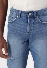 Muž ve světlých modrých džínách s mírným roztržením blízko levého stehna a s košilí béžové barvy zasunutou do kalhot.