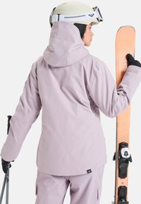 Veste de ski violet clair avec capuche, présentant une texture lisse. La figure tient un ski orange et porte un casque blanc. Gants noirs visibles.