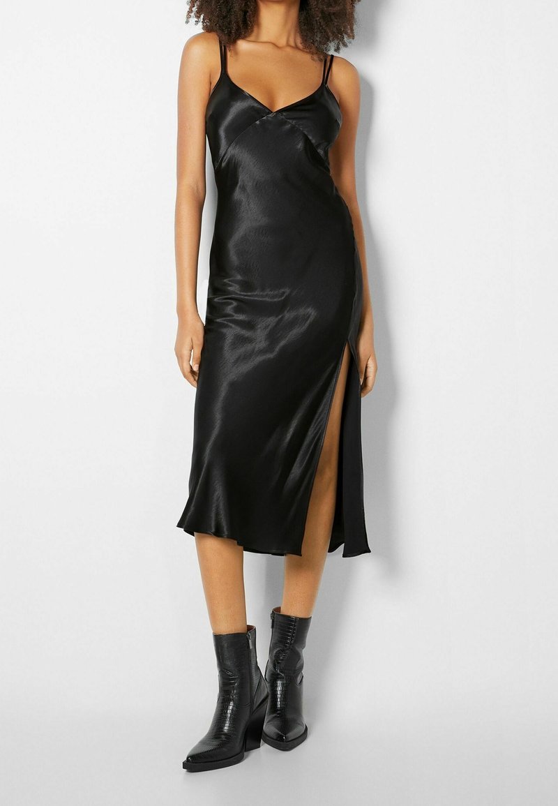 Femme portant une robe nuisette en satin noir à fines bretelles avec une fente haute, associée à des bottines noires mi-mollet à talons.