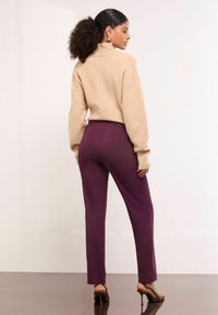 Pulover bej din tricot cu guler înalt și umeri căzuți, asortat cu pantaloni violeți cu talie înaltă și picioare drepte, și pantofi cu toc înalt de culoare maro.