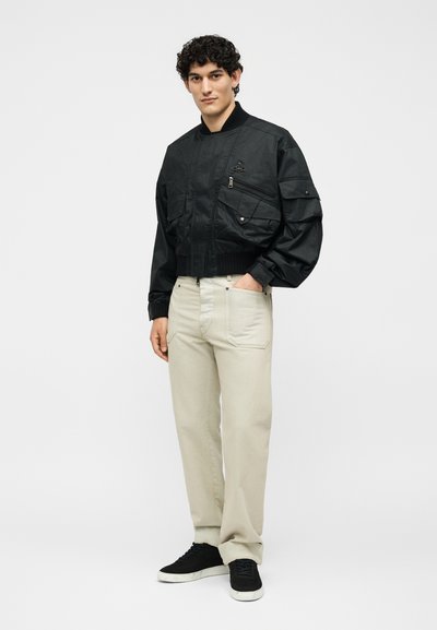 Άνδρας μοντέλο στέκεται με το χέρι στην τσέπη, φοράει μαύρο bomber μπουφάν με πολλές τσέπες, μπεζ παντελόνι και μαύρα αθλητικά παπούτσια με λευκές σόλες.