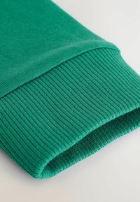 Puño de sudadera verde con textura acanalada, tejido superior liso y borde doblado. El material parece suave y ligero.
