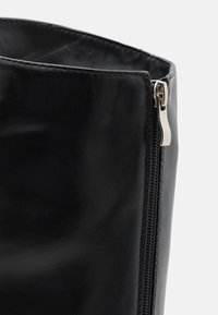 Gros plan sur la tige d'une botte en cuir noir, mettant en avant une fermeture éclair latérale en métal argenté partiellement ouverte, montrant la doublure intérieure.