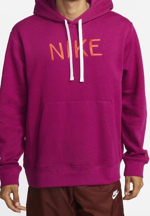 Mann trägt leuchtend lila Nike-Hoodie mit weißen Kordeln und braune Nike-Hose, steht vor einfachem Hintergrund.