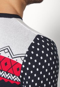 Gros plan sur une personne portant un pull en tricot avec des motifs géométriques en rouge, blanc et bleu marine, comprenant de petits carrés blancs sur la manche.