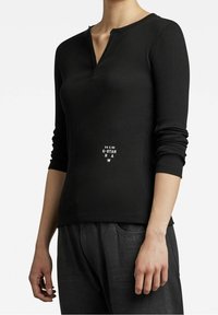 Schwarzes langärmliges Rippenoberteil mit V-Ausschnitt, ausgestattet mit einem Logo-Design auf der Vorderseite. Das Material wirkt weich mit einer leichten Textur und ist figurbetont.