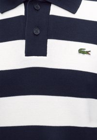 Polo en maille à rayures bleu marine et blanc avec deux boutons et petit logo crocodile vert sur la poitrine.