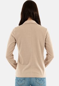 Camicia beige a maniche lunghe con tessuto testurizzato, dotata di colletto a scollo e dettaglio della cucitura sul retro, indossata con jeans in denim blu chiaro.