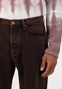Jeans de mezclilla marrón con un corte clásico, con cierre de botones en la parte frontal, detalles de costura sutiles y bolsillos laterales. Suéter en tonos claros arriba.