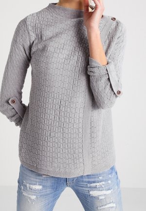 Sweater gris texturé avec un col haut et des accents de boutons sur les manches retroussées, associé à un jean bleu déchiré.