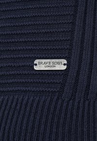 Tissu tricoté marine avec une texture côtelée horizontale et verticale. Comprend une étiquette en métal avec l'inscription "BRAVE SOUL LONDON" en relief.