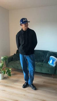 Black button-up jacket, blue jeans, and black shoes; wearing a blue Dodgers cap. Standing in front of a green couch with decorative pillow.