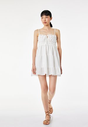 Femme portant une mini-robe blanche sans manches à volants et des sandales marron à brides, debout sur un fond blanc uni.