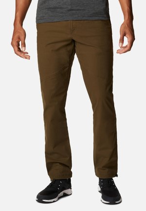 Mann trägt braune gerade geschnittene Hose und schwarze Turnschuhe, steht mit entspannten Händen an den Seiten vor weißem Hintergrund.