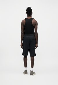 Άνδρας που στέκεται με την πλάτη γυρισμένη, φοράει μαύρο αμάνικο μπλουζάκι, μαύρο σορτς με πίσω τσέπη, λευκές κάλτσες και γκρι παπούτσια υψηλού πάνω μέρους.