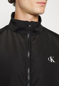 Czarna kurtka na zamek z wysokim kołnierzem, o gładkiej fakturze i białym logo "cK" na piersi, minimalistyczny design i elegancka sylwetka.