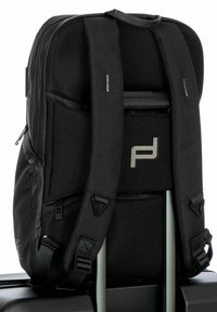 Mochila negra con espalda y correas de malla acolchada, que cuenta con bolsillos con cremallera y un logo de Porsche Design. Diseñada para sujetarse al equipaje.