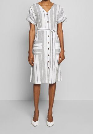 Kobieta ubrana w białą sukienkę o długości do kolan z pionowymi szarymi paskami, krótkimi podwiniętymi rękawami, guzikami z przodu, paskiem wiązanym w talii oraz białe, spiczaste buty.
