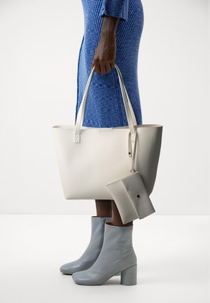 Personne en robe bleu côtelée tenant un sac fourre-tout écru avec une pochette assortie, portant des bottines gris clair à talons blocs.
