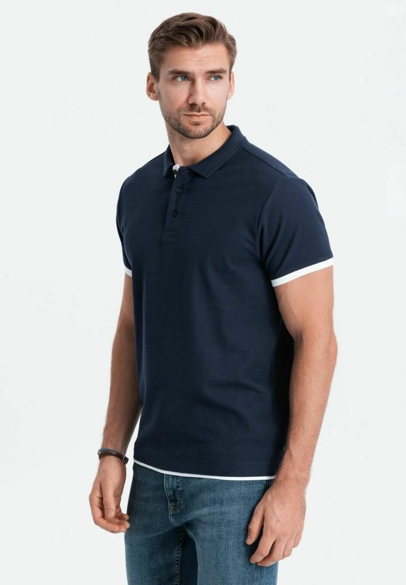 Marinblå polo med kort ärm, vita detaljer vid ärmsluten och treknapplås, matchad med mörka jeans. Slät textur.