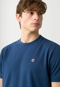 Mörkblå t-shirt i mjukt tyg, med rund halsringning och liten cirkulär logotyp på bröstet. Korta ärmar, figurnära design.