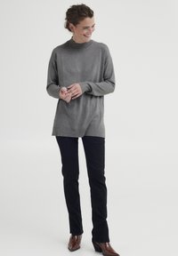 Graues Langarmoberteil mit hohem Kragen, kombiniert mit schwarzen Slim-Fit-Hosen. Das Model trägt burgunderfarbene spitze Stiefel und präsentiert einen lässigen Look.