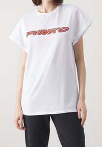 Bílý tričko s krátkým rukávem vyrobené z bavlny, s výrazným, stylizovaným logem "PINKO" v červené a černé barvě přes hrudník.