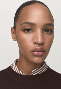 Young person with smooth skin and pulled-back hair wearing a brown sweater over a striped shirt, neutral expression, plain background.