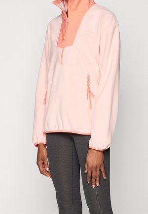 Osoba ubrana w jasnoróżową bluzę polarową z zamkiem do ćwiartki oraz zamkiem przy kieszeni bocznej, w połączeniu z ciemnoszarymi legginsami, stojąca na tle białego tła.