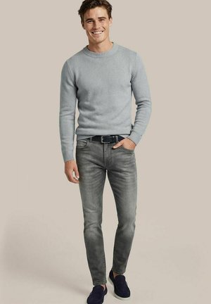 Junger Mann lächelt, trägt hellgrauen Pullover, ausgewaschene graue Jeans, schwarzen Gürtel und dunkle Slipper, steht vor einfarbigem Hintergrund.