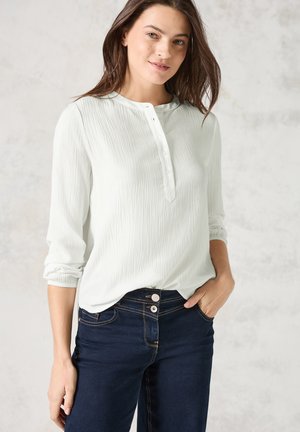 Blouse vert clair, texturée, avec un col henley et une fermeture à boutons, associée à un jean bleu foncé avec une fermeture à boutons visible.