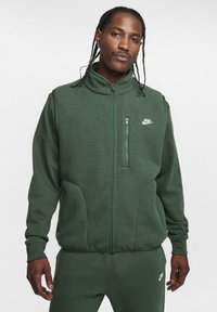 Colete de fleece verde com fecho de correr na frente, gola alta e bolsos laterais. Apresenta um pequeno logotipo branco da Nike no peito.