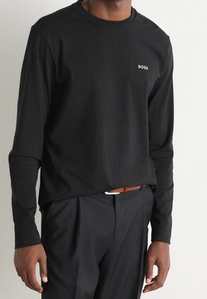Homme portant une chemise noire à manches longues avec un petit logo beige "BOSS" sur la poitrine, associée à un pantalon foncé et une ceinture marron.