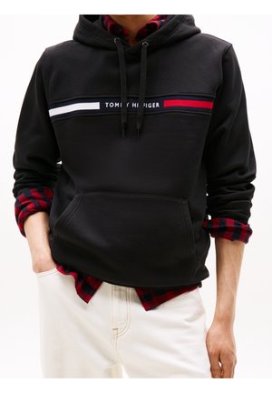Osoba nosząca czarną bluzę z kapturem Tommy Hilfiger na czerwono-czarnej koszuli w kratę, w połączeniu z białymi spodniami, ręce w kieszeniach.
