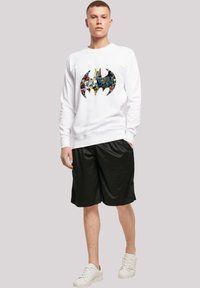 Hvid sweatshirt med et stort, flerfarvet Batman-logo, parret med sorte shorts. Modellen har hvide sneakers på.