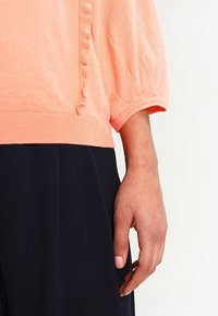 Broskvový pletený svetr s nafouknutými krátkými rukávy, zdobený texturovaným volánem po straně. Kombinován s tmavou plisovanou sukní.