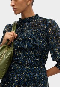 Czarna sukienka w kwiaty z niebieskimi i żółtymi kwiatami, zebrana faktura, wysoka szyja i puffowe rękawy; zestawiona z zieloną skórzaną torebką.