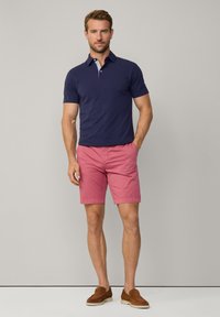 Polo shirt blu navy con maniche corte e colletto, abbinato a pantaloni corti rosa chiaro. Uomo in piedi con scarpe slip-on marroni, sfondo neutro.