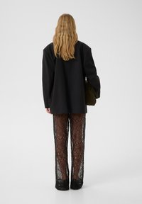 Svart oversized blazer parat med genomskinliga svarta spetsbyxor som har intrikata mönster och svarta skor. Modellen bär en olivgrön väska.