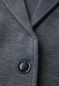 Šedý blazer s texturovanou tkaninou, klopou a tmavým knoflíkem s čtyřmi otvory. Záběr na vzor materiálu a detaily knoflíku.